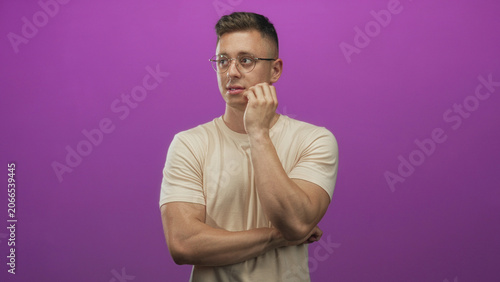 Canvas Print Young man with glasses biting nails, hand to mouth, beige tshirt against purple backdrop in studio; nervousness hesitation doubt