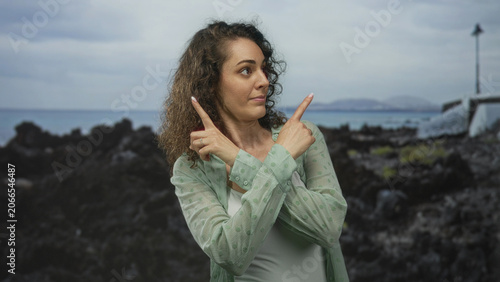 Fotografie Woman pointing finger to sky at rocky beach by the sea, wearing white camisole and sheer green shirt; calm contemplation