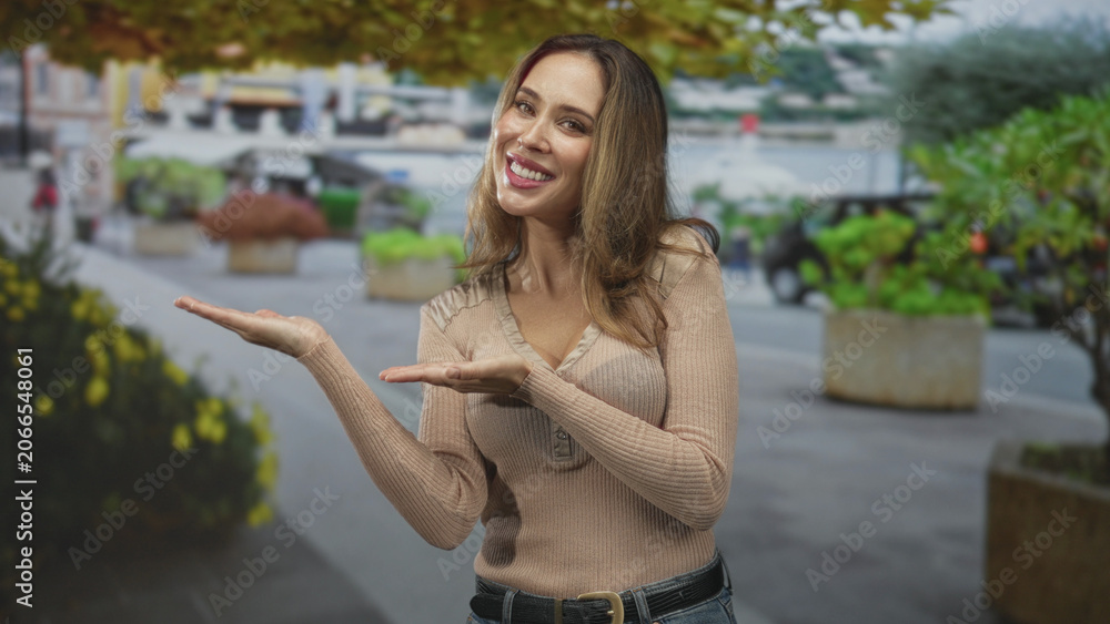 Fototapeta premium Woman palms up presenting with open hands and smiling on a city street beside planters and walkway; friendly welcome.