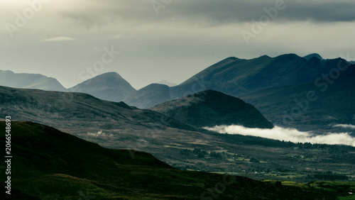 Low-lying clouds and mist drift through a valley dotted with fields and trees, surrounded by dark mountains.