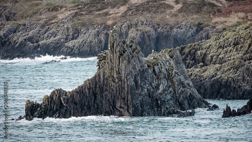 Jagged rock formations rise from the ocean, with waves crashing against their base and a dry, grassy hillside in the background.
