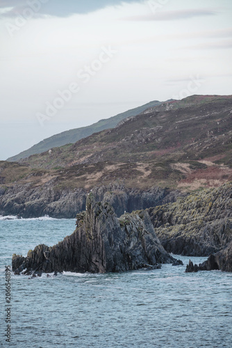 Jagged dark gray rock formations rise from the blue ocean water near a moss-covered rocky shore.