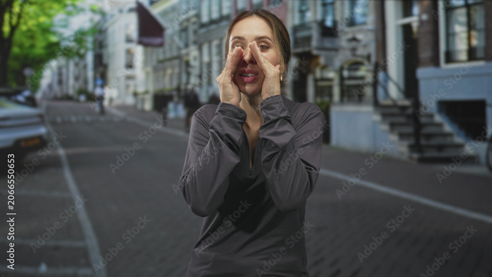 Fototapeta premium Woman cupping hands around mouth shouting to call attention on a city street with row houses and parked car; urgency warning.