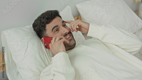 Foto Man lying in bed holding smartphone to ear, smiling and gesturing with hand in a bedroom; relaxed conversation