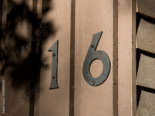 Forged Iron House Number 16 on an Old Wooden Cottage Wall. Evening Sunlight Creates Soft Leaf Shadows on Rustic Timber Cladding
