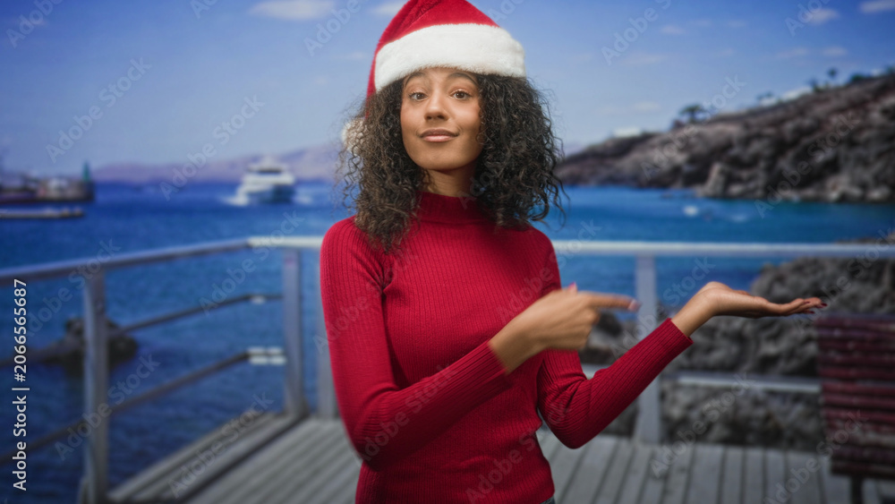 Fototapeta premium Woman points finger to open palm while wearing a christmas hat on a studio dock set by the sea; festive cheer.