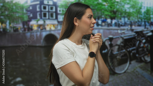 Schilderij op canvas Woman brunette clasping hands on street beside amsterdam canal, wearing white t shirt and smartwatch, looking thoughtful; reflection hope serenity