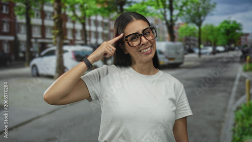 Woman wearing large glasses and white t shirt points finger to head on a city street with parked cars and trees visible; playful idea confidence.