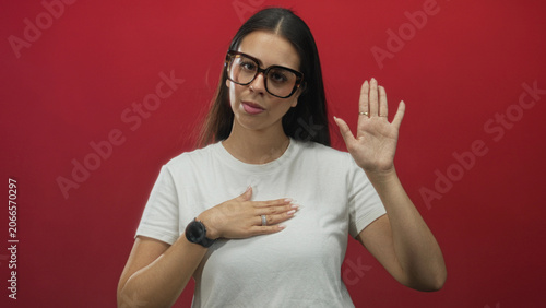 Fotografie Woman with hand on chest and raised hand wearing glasses and white t shirt, showing rings and watch, in red studio; sincerity commitment