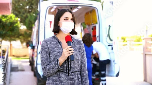 A woman wearing a face mask holds a microphone while medical staff transport a patient on a stretcher from an ambulance, conveying a sense of breaking news in the healthcare or emergency services fiel