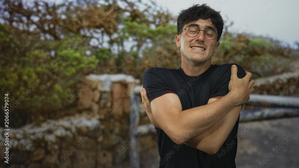 Fototapeta premium Man smiling with arms crossed hugging himself in a forest setting by stone wall, wearing glasses and black shirt; comfort selfcare.