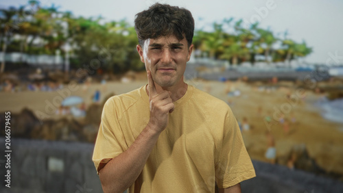 Photos Man in yellow t shirt finger to cheek for toothache on crowded sandy beach by palm trees and seawall, squinting in sunlight; discomfort