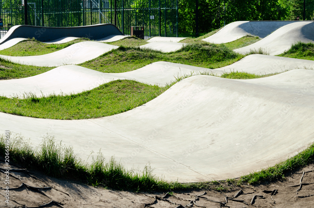 Naklejka premium concrete bike track with wave ramps in park area. this track provides challenging course for cycling and bmx riding. modern track blends with green grass and trees for outdoor recreation.