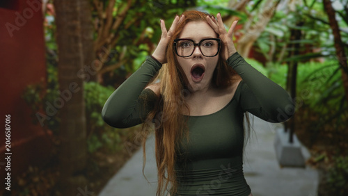 Obraz na plátně Redhead woman wearing oversized glasses and off-shoulder green top holds hands to head with mouth agape on a forest path lined with tropical foliage and concrete walkway; surprise discovery