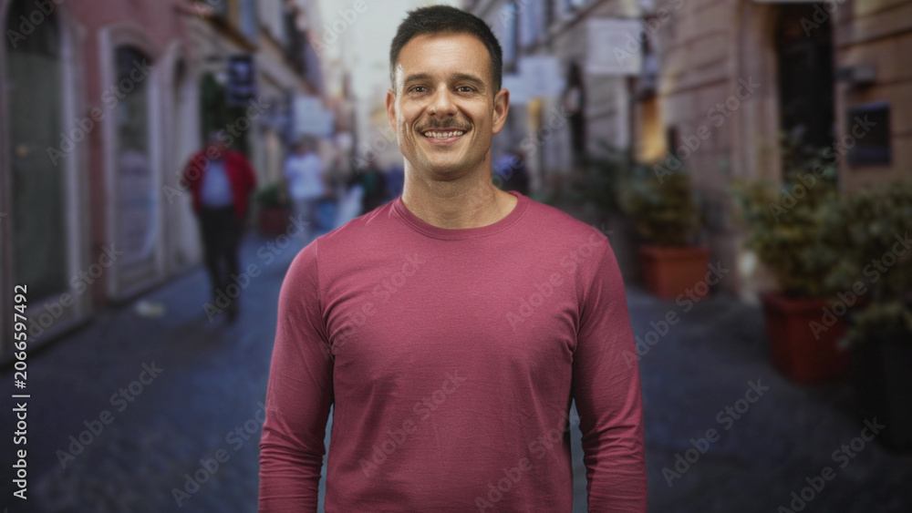 Obraz premium Man with mustache turning head and smiling while wearing a crewneck shirt on a narrow cobblestone street lined with shops; friendly confidence.