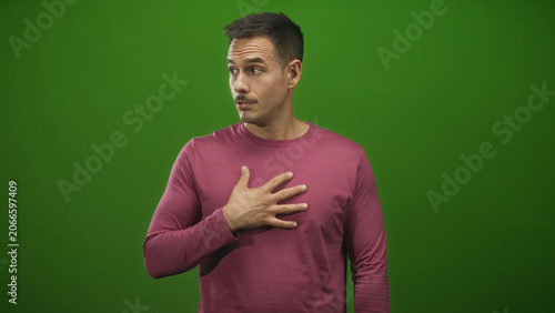 Obraz na plátně Man with right hand on chest and pursed lips in studio with green backdrop, glance sideways and hesitant posture suggesting self doubt and surprise; vulnerability surprise