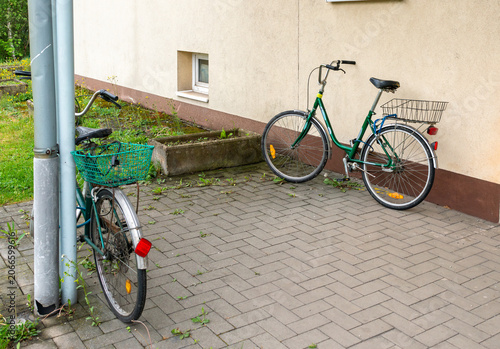 Two classic utility bicycles with baskets parked next to a residential building wall. The composition illustrates sustainable transportation, everyday cycling and urban neighborhood life.