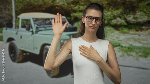 Фотография Woman raises hand with other hand on chest beside a green offroad vehicle in forest wearing white sleeveless garment; oath sincerity