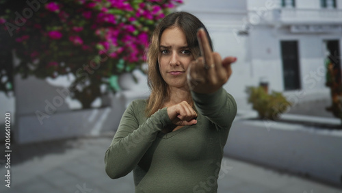 Slika na platnu Young hispanic woman shows middle finger to camera in a city street framed by white building and pink bougainvillea; defiance