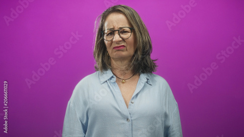 Fotografía Woman, senior with grey hair, frowning, wearing glasses and pendant necklace, standing in a purple studio; displeasure reflection