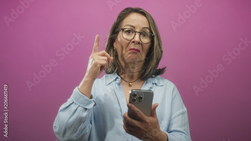 Bild auf Leinwand Woman pointing finger while holding smartphone in studio with pink backdrop, wearing glasses and necklace; disapproval warning