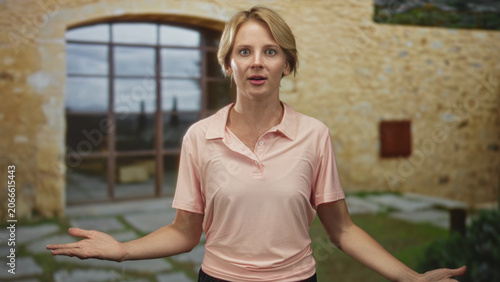 Tableau sur toile Woman wearing pink polo shirt, hands open, speaking and gesturing in front of a stone building window and courtyard; calm explanation