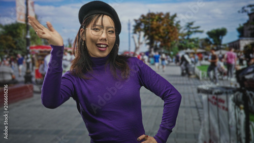 Cuadro en lienzo Woman wearing beret and glasses waving hand and smiling with ringed fingers in a purple sweater on a busy street market with food stalls and crowds; friendly joy