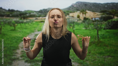 Bilde på lerret Woman holding two pairs of glasses and puckering lips while testing frames in front of building; playful choice