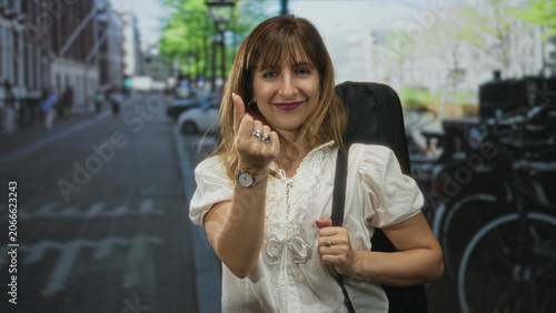 Photos Woman beckons with hand and fingers while holding a guitar case on a busy city street lined with bicycles and parked cars; playful