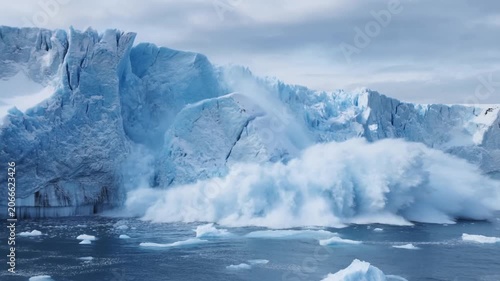 Slow motion tracking shot of glacier calving and massive iceberg collapse into arctic ocean seascape with splashing waves and falling ice