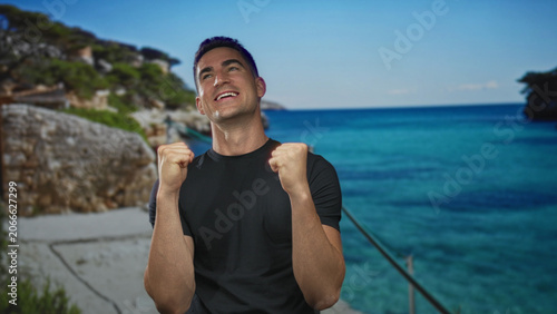 Valokuva Man with clenched fists on a sunny seaside street near rocky cliffs and turquoise water; elation victory