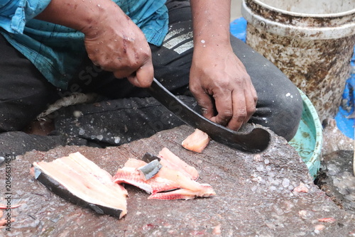  person in a blue shirt is filleting fish using a large, curved knife and  fish scales and small pieces scattered around