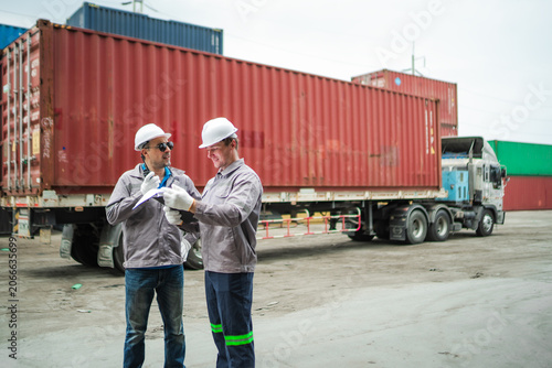 Logistics workers review shipment verification documents beside cargo truck and shipping container, coordinating freight logistics, transportation planning, and supply chain operations.