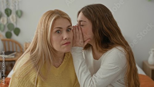 Fotografía Blonde mother listens as her daughter whispers into her ear with hand covering mouth while seated on a bed in a bedroom; surprise intimacy trust