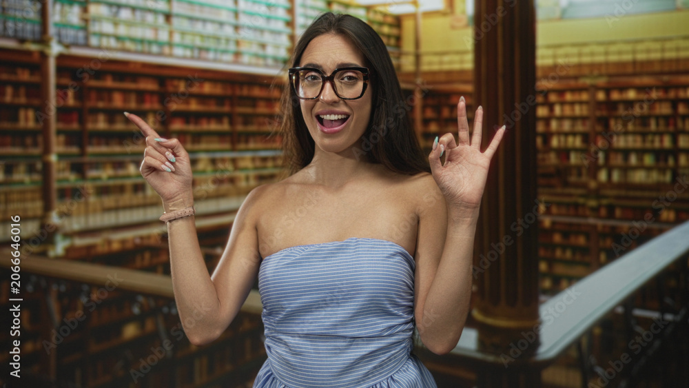 Fototapeta premium Woman points finger and makes ok sign with hand in ornate library among bookshelves, showing bare shoulders and glasses; approval study concentration.