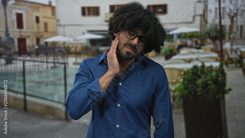 Tableau sur toile Man with curly hair and glasses touching cheek with hand for toothache at restaurant terrace; discomfort