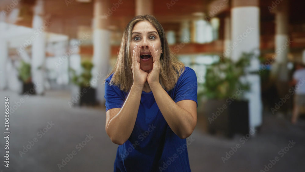 Fototapeta premium Woman cupping hands at mouth calling across a busy airport terminal near planters and white columns; urgent announcement.