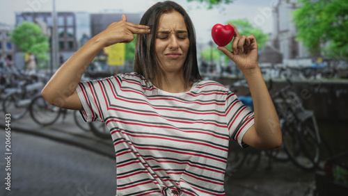 Konstfotografi Woman holding red heart and pointing two fingers upward on a street in amsterdam; love irony sarcasm
