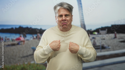 Foto Senior man grips sweater with both fists and points to his chest at a crowded beach with a visible palm tree backdrop; disgust