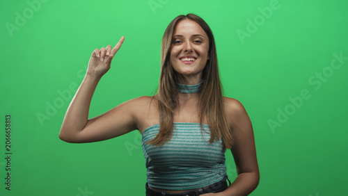 Fotografie Woman points index finger up in studio green screen while smiling, finger raised and arm slightly bent; confidence