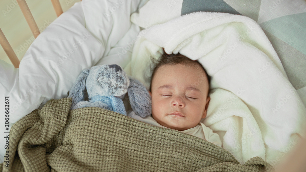 Fototapeta premium Baby boy sleeping with closed eyes in a crib, face visible, swaddled under a green blanket beside a plush rabbit inside a building; serene rest.