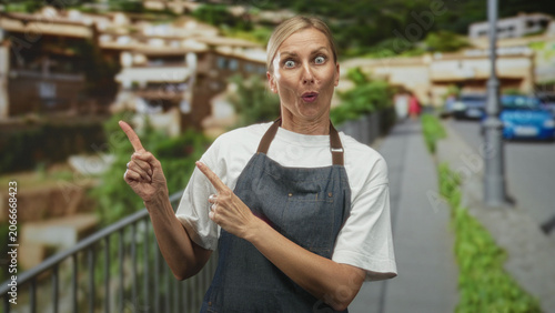 Canvas Print Middle age blonde woman in apron points finger with visible forefinger and surprised puckered lips on street; surprise curiosity