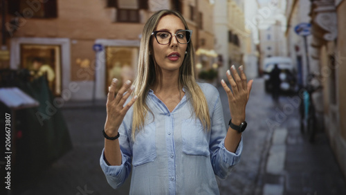 Photos Woman with hands raised making a speaking gesture on a city street, wearing glasses and light blue shirt; serenity