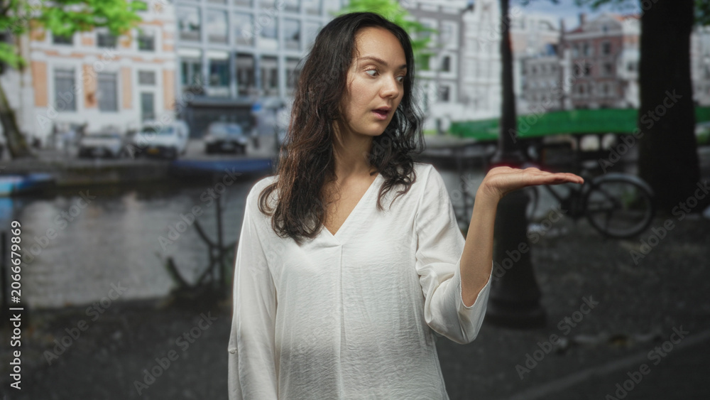 Fototapeta premium Woman in white blouse holds palm up, presenting on a street by a canal with bicycle and boats visible; curiosity.