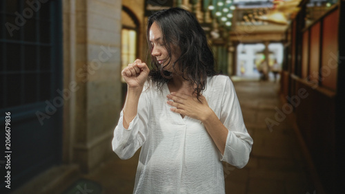 Foto Woman coughing into fist while her hand presses chest, wearing white shirt, standing in a building corridor; discomfort
