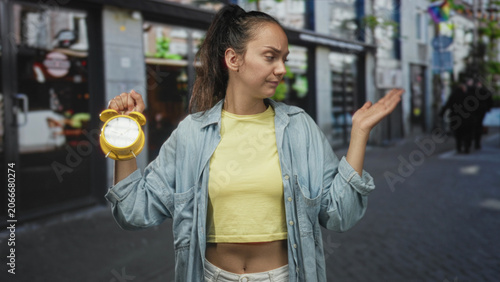 фотографія Young hispanic woman holding yellow alarm clock and shrugging one hand in a busy city street wearing yellow crop top and denim shirt; frustration time pressure