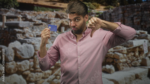 Fotografija Man holding creditcard while showing thumbs down gesture in building inspecting a payment card; disappointment caution