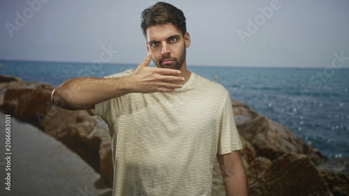 Fotografie Hispanic man covers mouth with hand and later touches forehead, standing on rocky beach near sea horizon in striped tee, direct gaze toward camera; silence