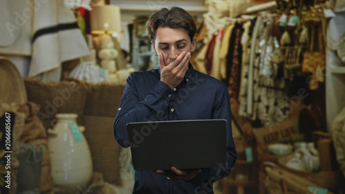 фотографія Man holding laptop with hand to mouth gesture inside a cluttered antique shop building; surprise worry