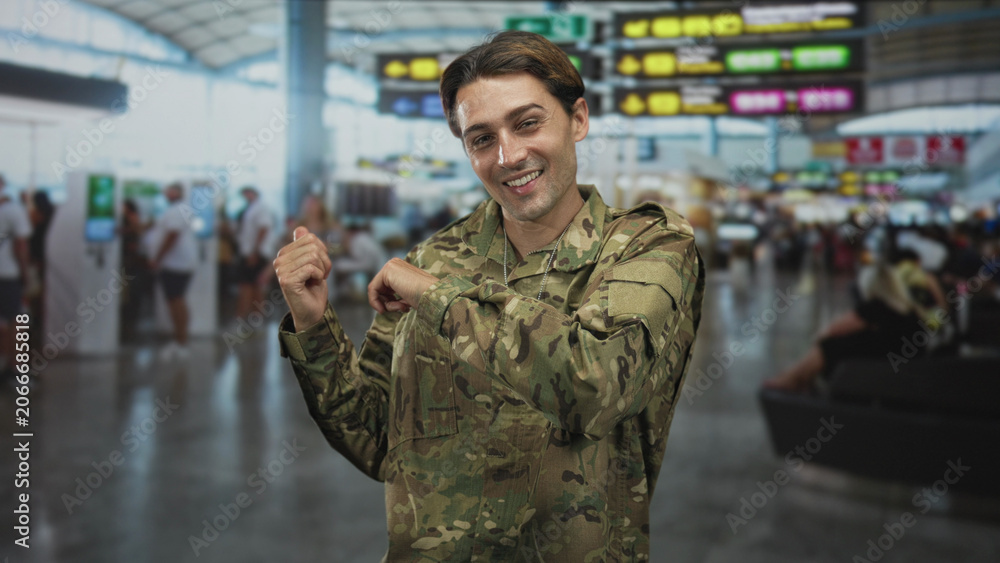 Fototapeta premium Soldier man in camouflage uniform adjusts dogtag and smiles toward camera in crowded airport terminal; pride reunion.